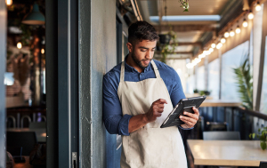 Young man in an apron using a tablet while standing in a modern restaurant, focused on managing orders or customer information. Bright interior with hanging lights and greenery in the background.