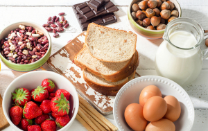 A vibrant assortment of healthy foods including fresh strawberries, eggs, whole grain bread, mixed beans, hazelnuts, dark chocolate, and a jug of milk, arranged on a rustic wooden surface.
