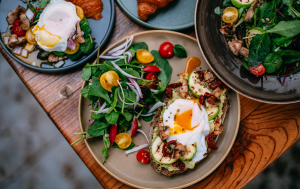 A beautifully arranged breakfast plate featuring avocado toast topped with a poached egg, grilled zucchini, sun-dried tomatoes, and sautéed mushrooms, accompanied by a fresh salad with cherry tomatoes and mixed greens. The setting includes a croissant on a separate plate, all presented on a rustic wooden table.