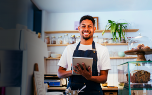 Smiling barista in a café holding a tablet, wearing a denim apron, with shelves of jars and baked goods in the background.